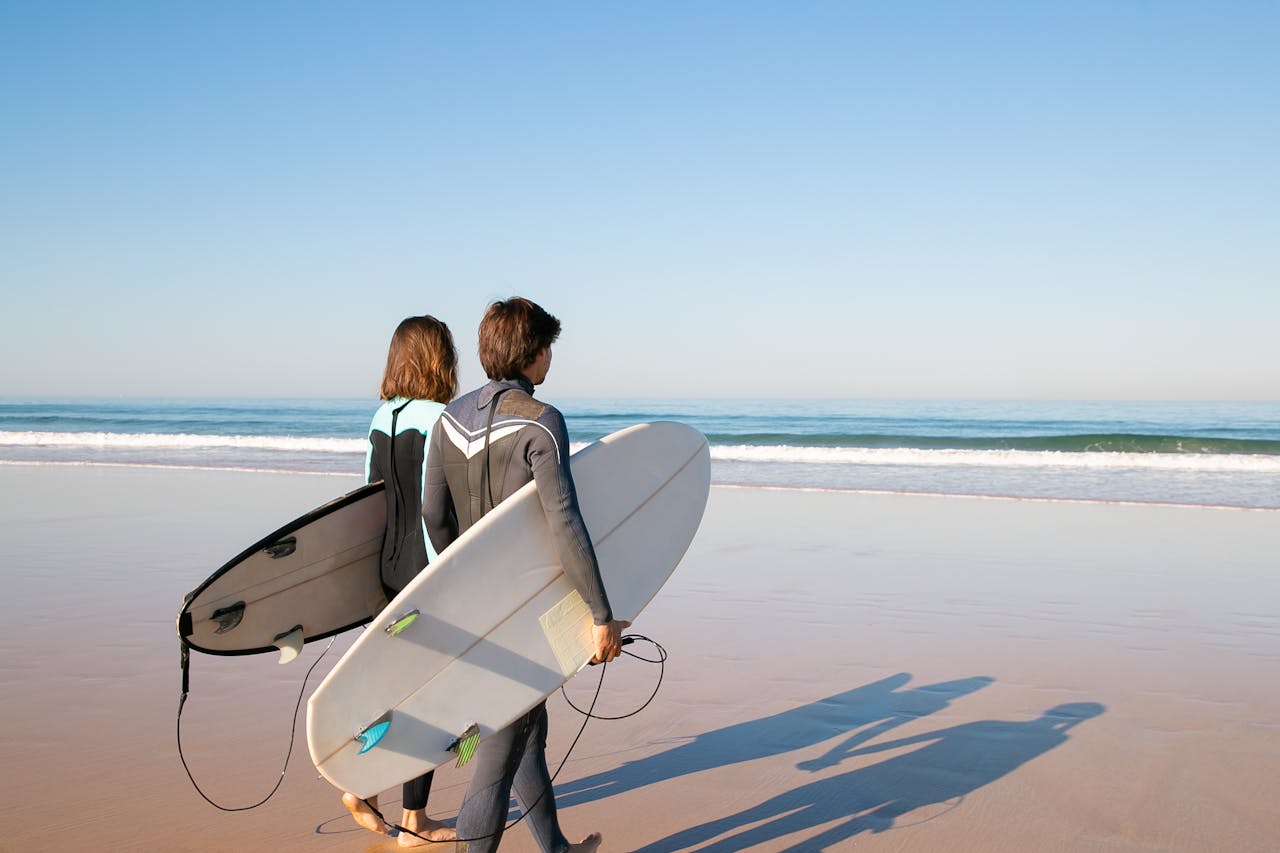 heros-img A young couple in wetsuits carrying surfboards along a sunny beach in Portugal.